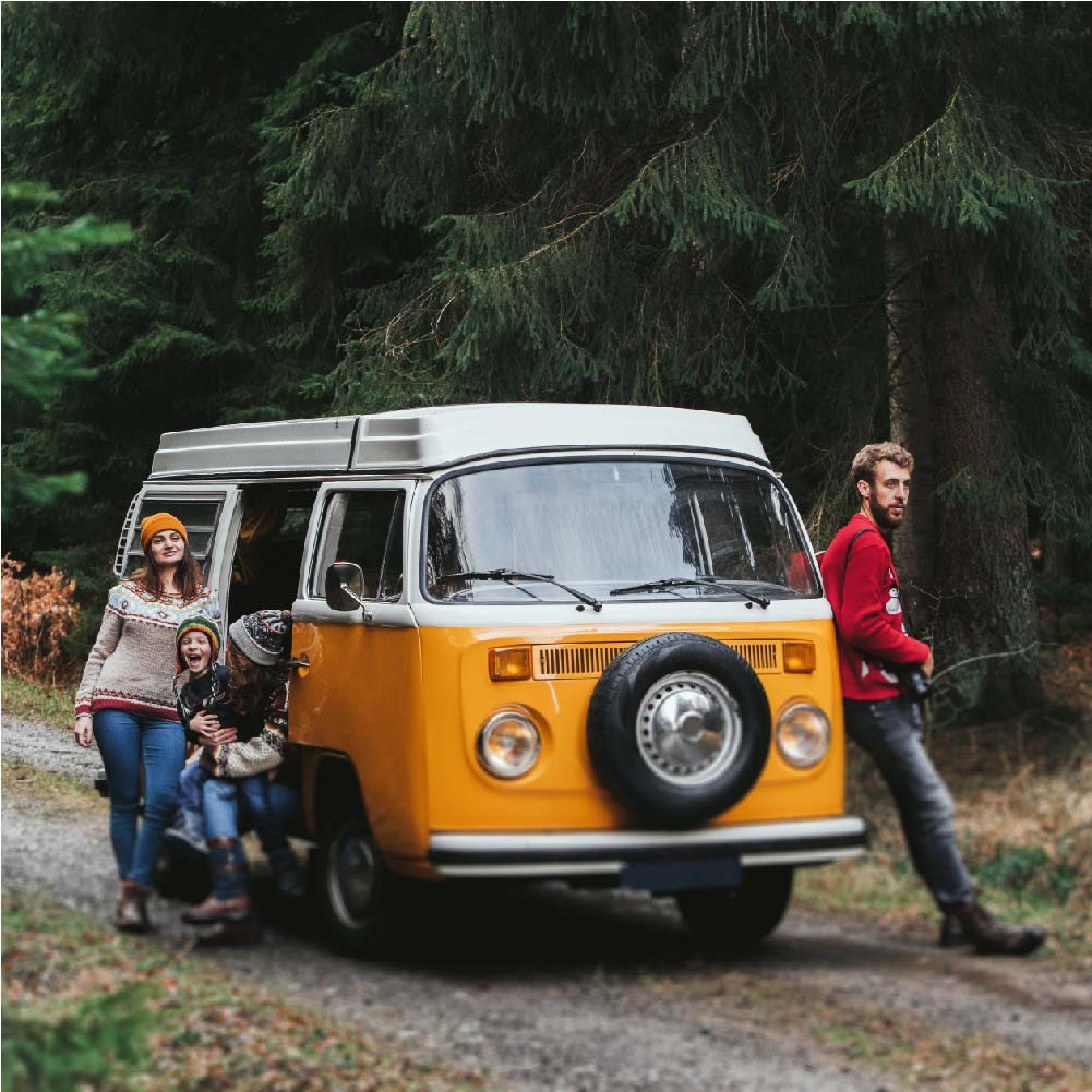 Image of a family happily leaning against their orange VW campervan, parked on a road surrounded by the tranquillity of a woodland setting.