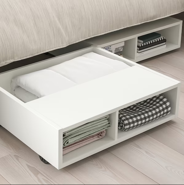 Partially open under-bed storage drawer filled with neatly folded sheets and books, highlighting efficient use of bedroom space.